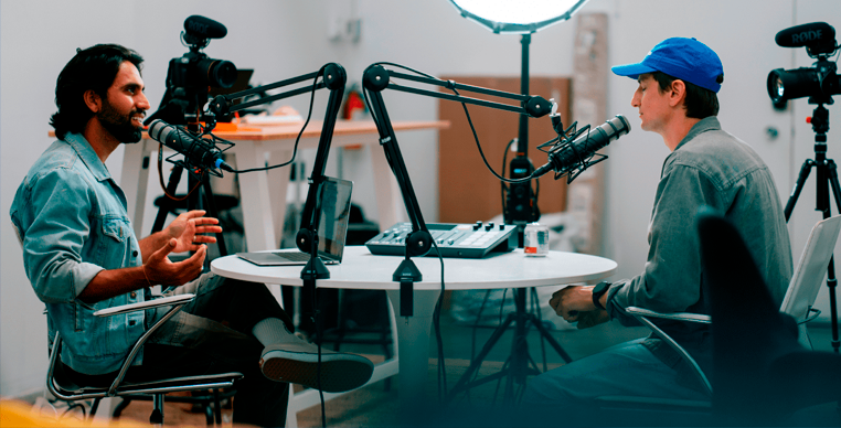 dois homens sentados em mesa com microfones gravando podcast